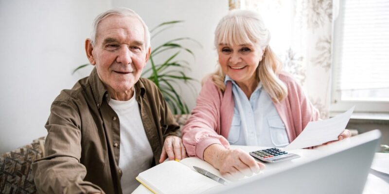 An older couple sitting at a table paying for senior living using a laptop.