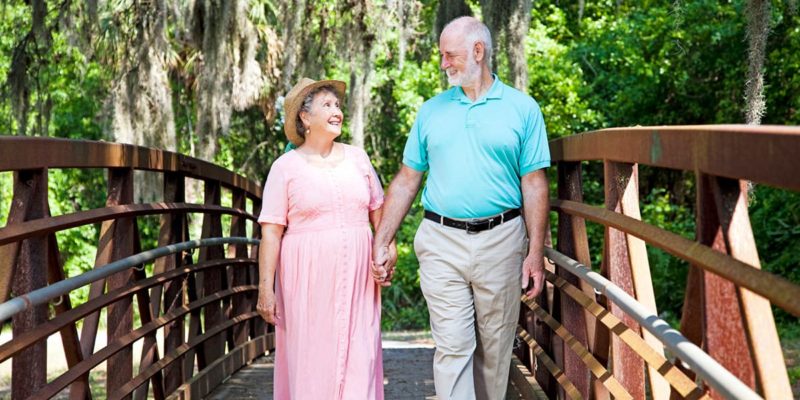 An older couple enjoying their retirement in the Tampa Bay area take a leisurely stroll on a bridge surrounded by the tranquil woods.