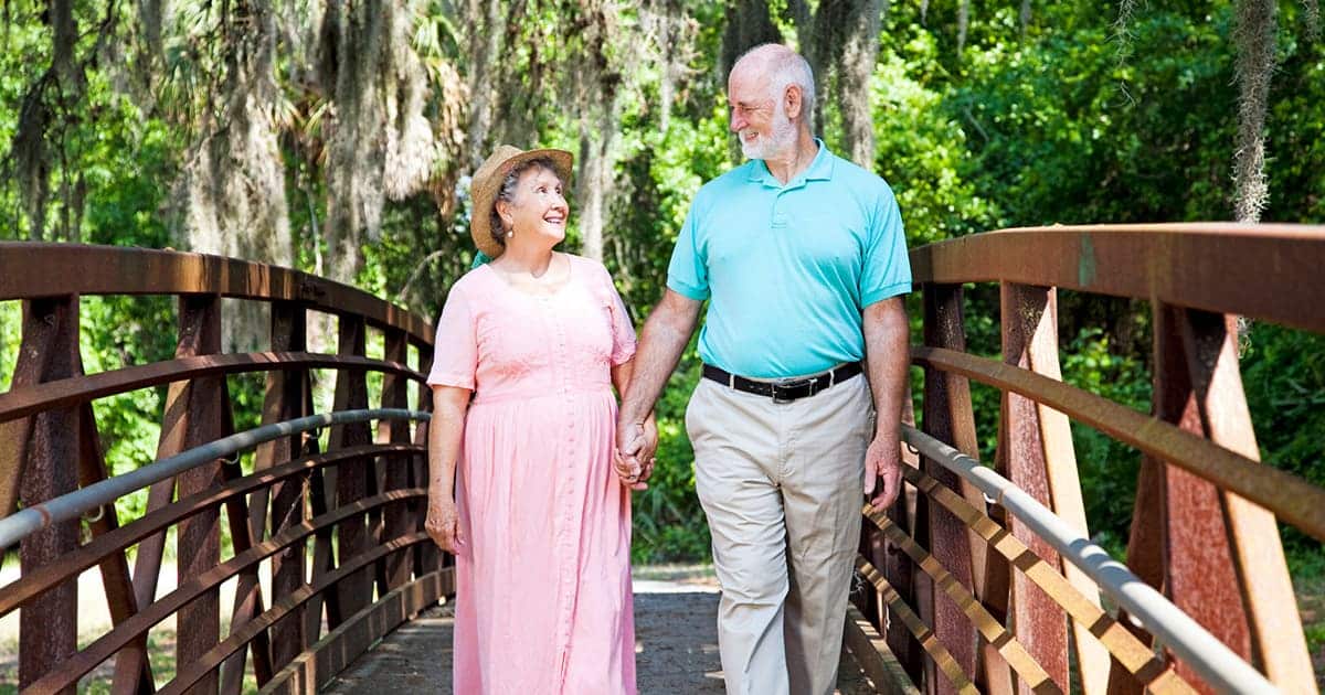 An older couple enjoying their retirement in the Tampa Bay area take a leisurely stroll on a bridge surrounded by the tranquil woods.