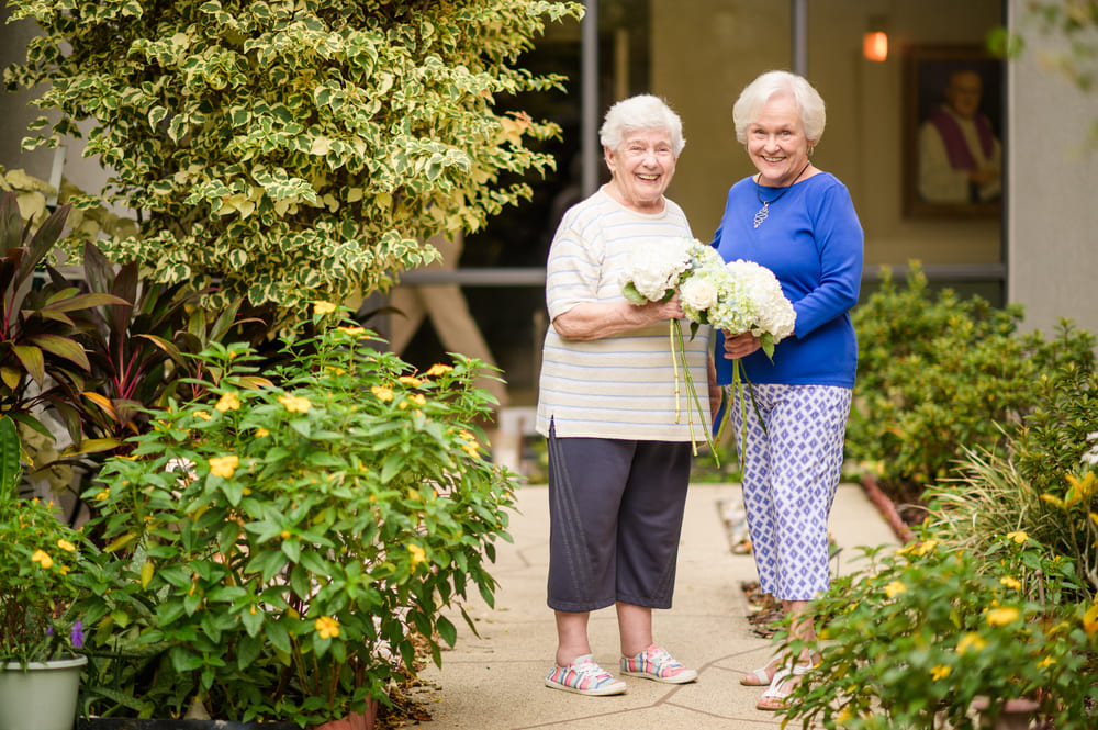 Two older women holding bouquets in front of a St Mark Village house.