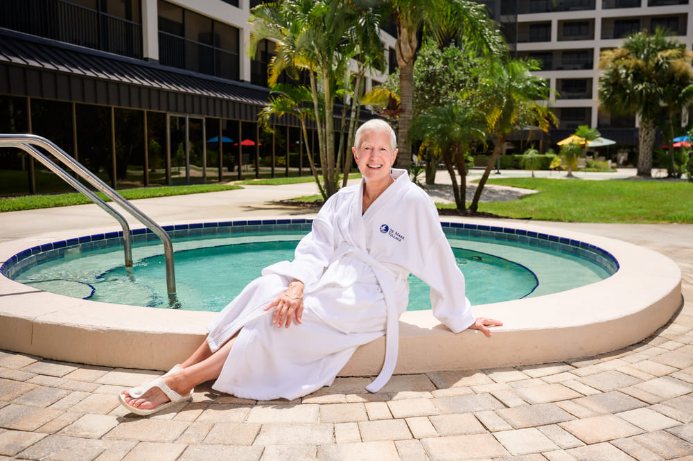 A woman in a white bathrobe enjoying the St Mark Village lifestyle by sitting in front of a pool.