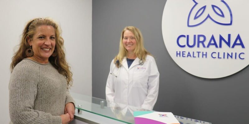 Two women standing in front of the curana health clinic sign.