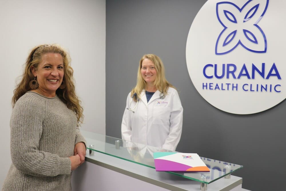 Two women standing in front of the curana health clinic sign.