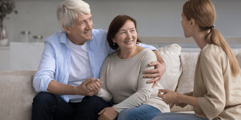 Senior couple sitting on sofa discussing senior living care options with adult female.