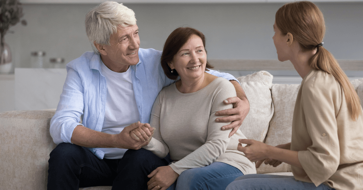 Senior couple sitting on sofa discussing senior living care options with adult female.