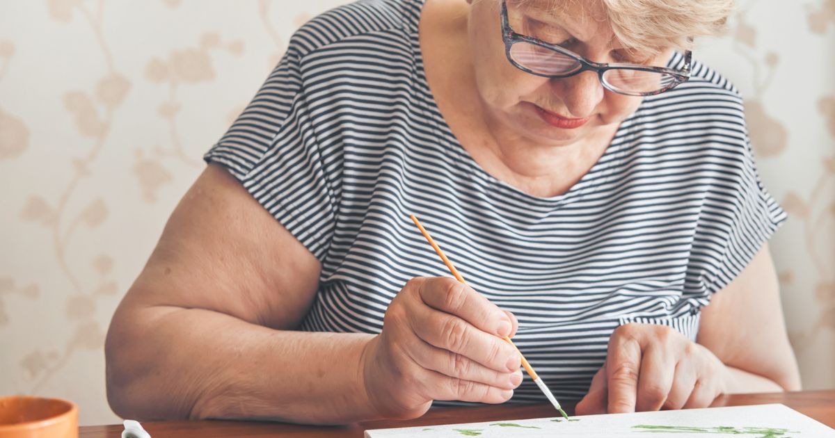 An older woman with glasses and a striped shirt is painting on paper with a paintbrush at a table, enjoying a creative afternoon at St. Mark Village senior living.