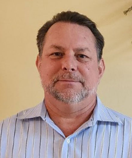 A middle-aged man with short brown hair and a beard, wearing a light blue striped collared shirt, stands in front of a plain light-colored wall at St. Mark Village senior living community. A middle-aged man with short brown hair and a beard, wearing a light blue striped collared shirt, stands in front of a plain light-colored wall at St. Mark Village senior living community.