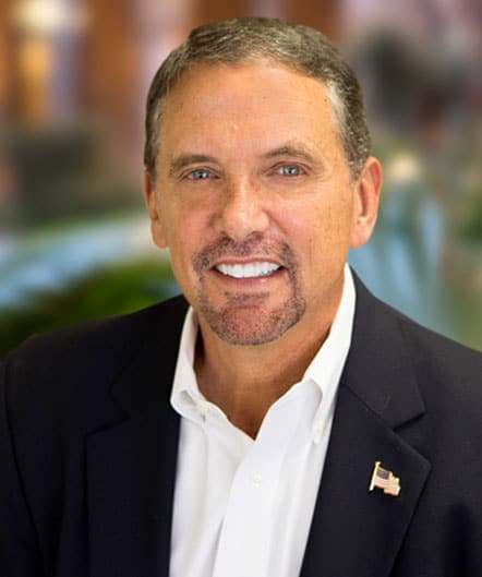 A man with short brown hair and a goatee, wearing a dark blazer, white shirt, and American flag pin, smiles at the camera with a blurred St. Mark Village senior living background. A man with short brown hair and a goatee, wearing a dark blazer, white shirt, and American flag pin, smiles at the camera with a blurred St. Mark Village senior living background.