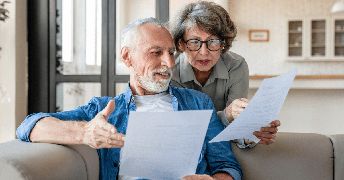 An older man and woman sit on a couch in a well-lit living room at St. Mark Village, looking at and discussing documents together, showcasing the vibrant senior living community.