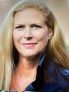 A woman with long blonde hair and a black collared jacket looks at the camera with a neutral expression against a blurred background, capturing a quiet moment at St. Mark Village senior living.