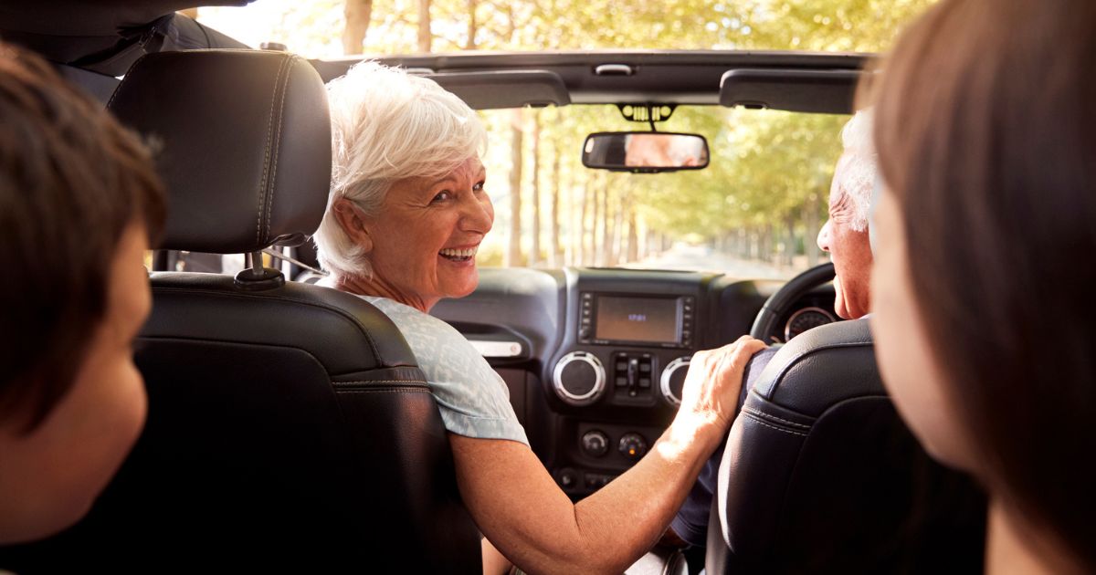 Older woman smiling in a car, sitting in the front passenger seat, looking back at two people in the back seat while another person drives through a sunlit area—enjoying senior living with friends from St. Mark Village.