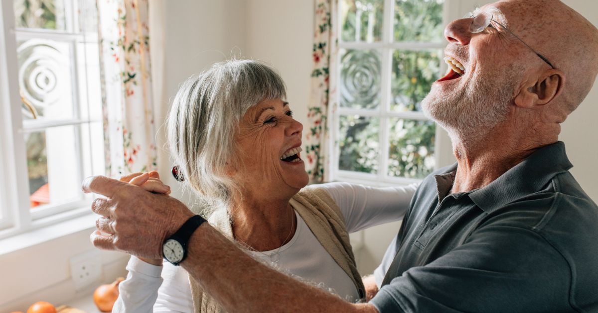 An older couple smiles and laughs while dancing together in a sunlit room with floral curtains, enjoying the vibrant community of St. Mark Village senior living.