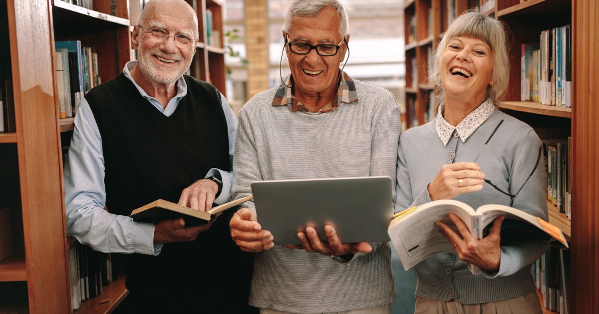 Three older adults stand in a library aisle at St. Mark Village senior living, holding books and a laptop, smiling and looking at the camera.