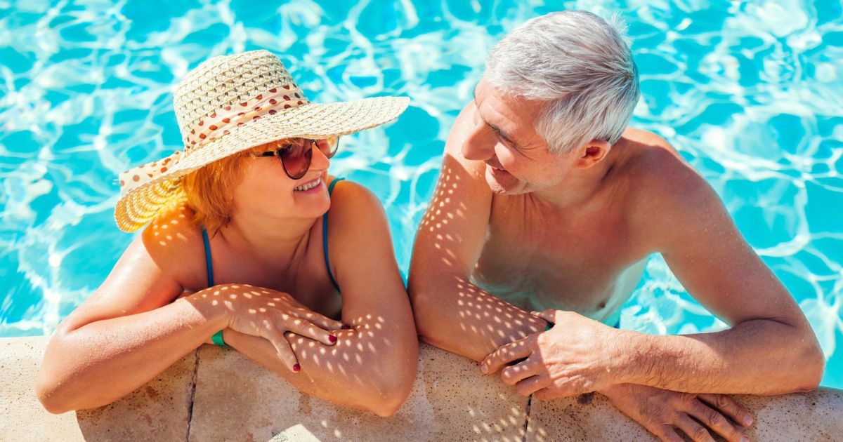 An older man and woman are smiling and talking while relaxing at the edge of a swimming pool in bright sunlight, enjoying the vibrant community atmosphere at St. Mark Village senior living.