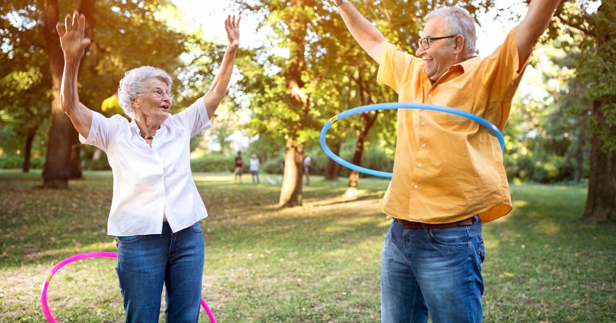 Two older adults, residents of St. Mark Village senior living, enjoy hula hooping—one with a pink and the other with a blue hoop—in a sunny park surrounded by trees and grass.