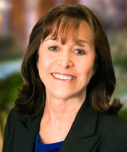 A woman with shoulder-length brown hair, wearing a black blazer and blue top, smiles at the camera against a blurred background at St. Mark Village senior living community. A woman with shoulder-length brown hair, wearing a black blazer and blue top, smiles at the camera against a blurred background at St. Mark Village senior living community.