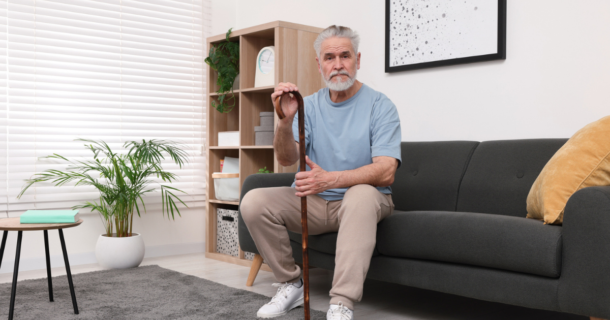 An older man with a beard sits on a sofa in a senior living community, holding a wooden cane and looking at the camera.