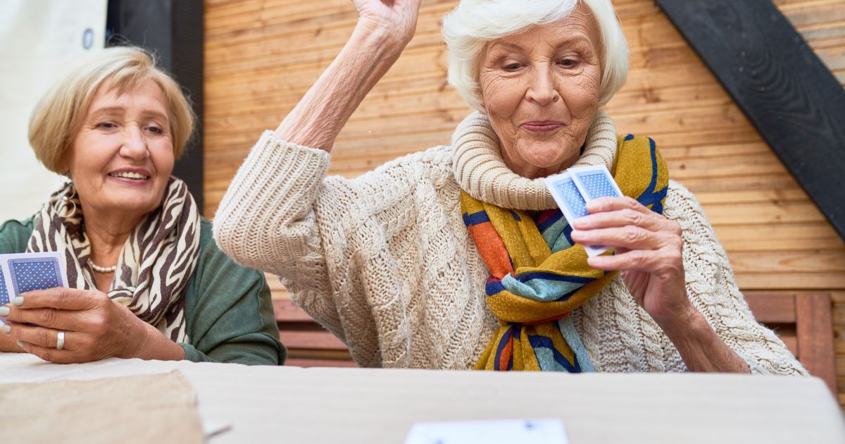 At St. Mark Village, two elderly women sit at a table playing cards. One woman holds her cards and raises her hand as if about to play, while the other smiles and watches, enjoying the vibrant senior living community.