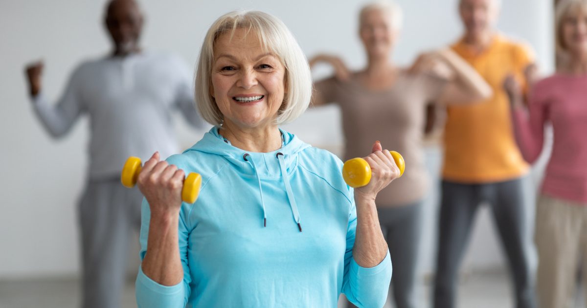 Older adults enjoying fitness together indoors at St. Mark Village senior living; a smiling woman in front holds yellow dumbbells while others pose in the background.