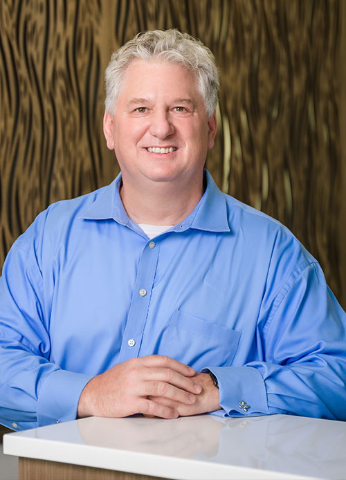 A middle-aged man with gray hair wearing a blue button-down shirt sits at a white counter, smiling at the camera. The warm, patterned brown wall behind him reflects the welcoming atmosphere of St. Mark Village senior living. A middle-aged man with gray hair wearing a blue button-down shirt sits at a white counter, smiling at the camera. The warm, patterned brown wall behind him reflects the welcoming atmosphere of St. Mark Village senior living.