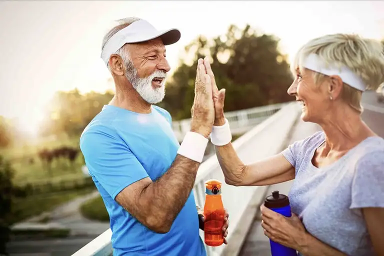 Two older adults in athletic wear high-five while holding water bottles outdoors on a sunny day at St. Mark Village, both smiling and wearing visors, enjoying the vibrant senior living community. Two older adults in athletic wear high-five while holding water bottles outdoors on a sunny day at St. Mark Village, both smiling and wearing visors, enjoying the vibrant senior living community.