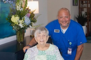 An elderly woman in a hospital gown sits in a chair while a smiling St. Mark Village healthcare worker in blue scrubs stands beside her. A vase of flowers adds warmth to this caring senior living environment.