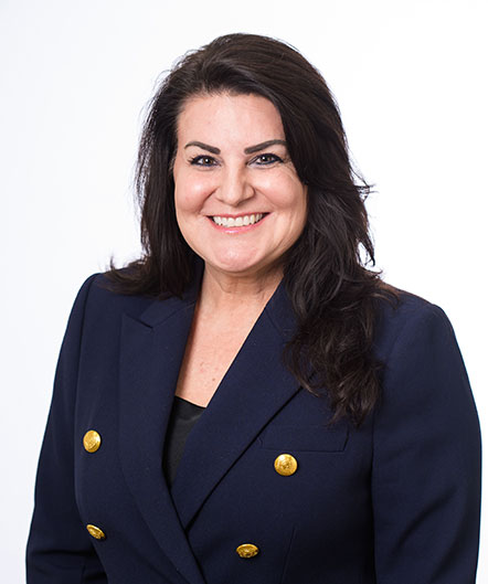 Woman with long dark hair wearing a navy blazer with gold buttons, smiling, in front of a plain white background, representing the welcoming spirit of St. Mark Village senior living. Woman with long dark hair wearing a navy blazer with gold buttons, smiling, in front of a plain white background, representing the welcoming spirit of St. Mark Village senior living.