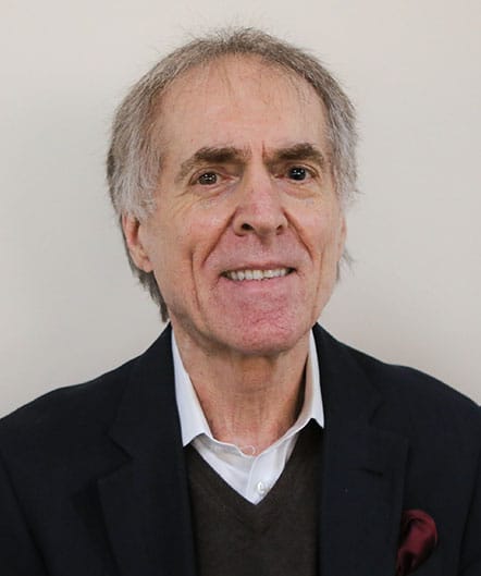 An older man with gray hair, wearing a dark blazer, white shirt, and brown sweater, smiles at the camera against a plain light background—reflecting the warmth and comfort found at St. Mark Village senior living. An older man with gray hair, wearing a dark blazer, white shirt, and brown sweater, smiles at the camera against a plain light background—reflecting the warmth and comfort found at St. Mark Village senior living.