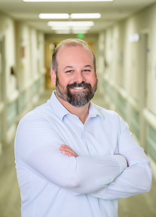 A man with a beard and mustache wearing a light blue shirt stands in a hallway at St. Mark Village, arms crossed and smiling at the camera, embodying the welcoming spirit of senior living.
