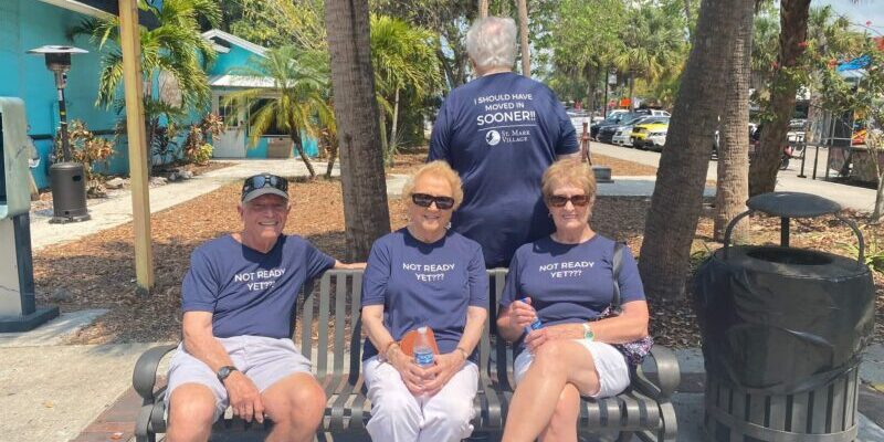 Four seniors wearing matching shirts sit and stand on a bench outdoors at St. Mark Village; three shirts say "NOT READY YET!!" and one says "I SHOULD HAVE MOVED IN SOONER," with palm trees in the background, celebrating senior living.