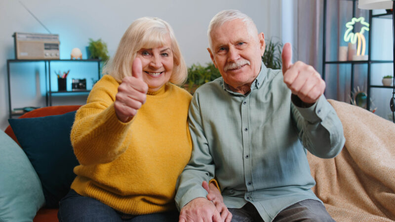 Couple on couch giving a thumbs up for how CCRCs protect seniors from increasing the costs of care