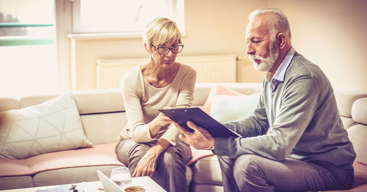 Two older adults sit on a couch at St. Mark Village, reviewing documents together; one holds a clipboard while the other looks on attentively, embracing the supportive environment of senior living.