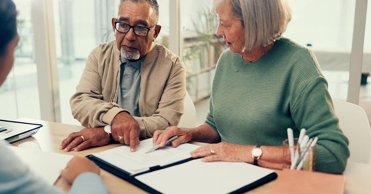 Two older adults review documents with another person at a desk, discussing paperwork in a bright, modern office setting at St. Mark Village senior living community.