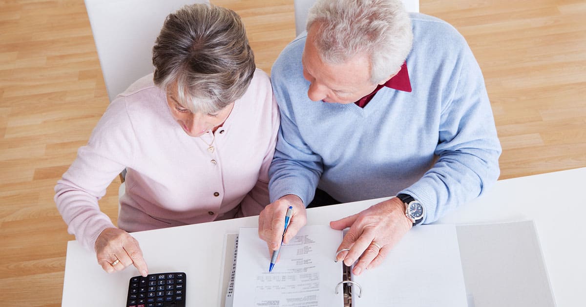 An older couple sits at a table in St. Mark Village senior living, reviewing documents—one using a calculator, the other holding a pen over a binder.