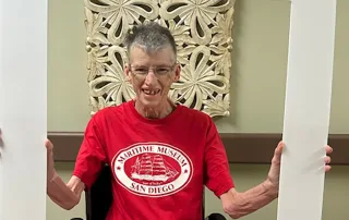 An older person in a red "Maritime Museum San Diego" shirt sits in a wheelchair, smiling and holding a "St. Mark Village - I DID IT!!!" photo frame, capturing the vibrant spirit of senior living.
