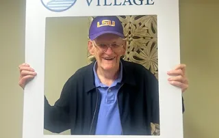 An elderly man in an LSU cap smiles at the camera, holding a large photo frame prop labeled "St. Mark Village" and "I DID IT!!!" to celebrate his achievement at a vibrant senior living community.