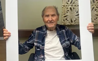An older man sits smiling inside a large white photo frame that reads “St. Mark Village” at the top and “I DID IT!!!” at the bottom, celebrating life in this vibrant senior living community.