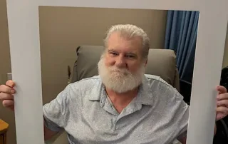 Older man with a white beard sits in a chair holding a large St. Mark Village senior living photo frame with the words "I DID IT!!!" at the bottom.