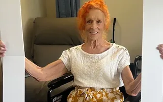 An elderly woman with orange hair sits in a wheelchair, smiling and holding a large frame that reads "St. Mark Village" and "I DID IT!!!," celebrating her achievements in senior living.