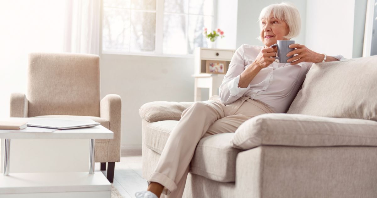 Older woman sitting on a sofa in a bright living room at St. Mark Village senior living, holding a mug and looking out the window, appearing relaxed.