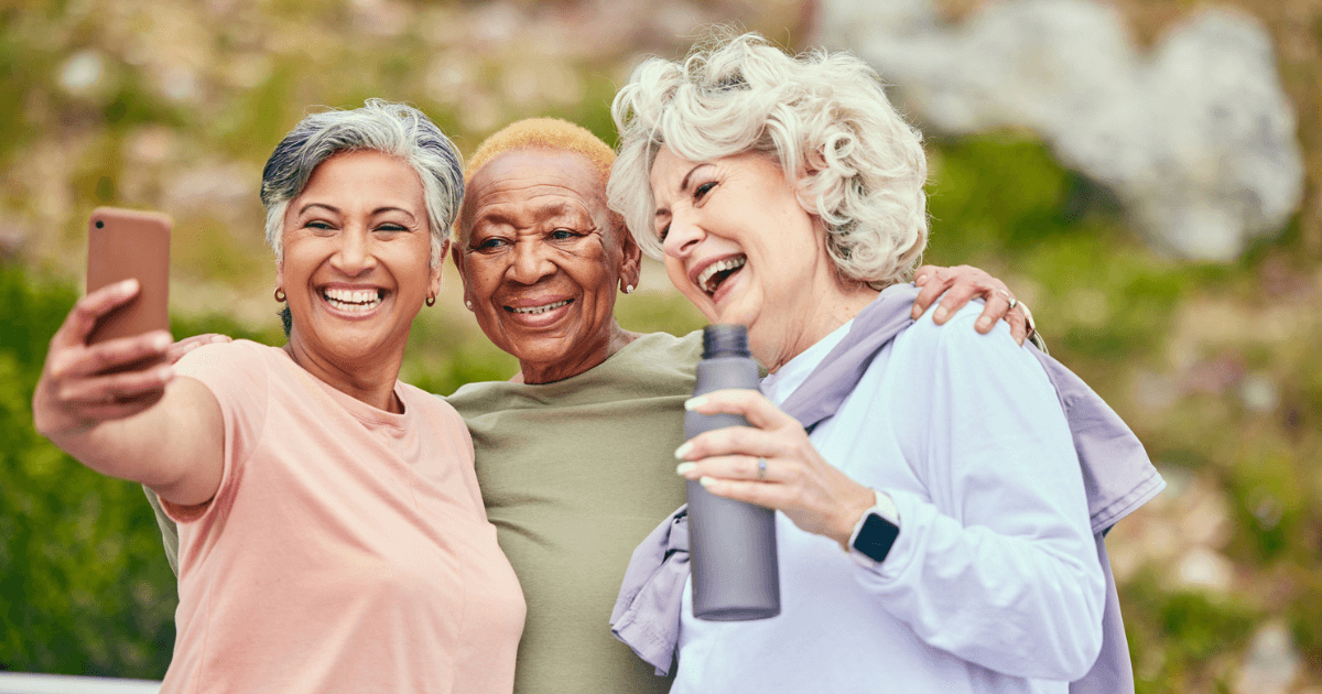 Three older women at St. Mark Village are smiling and posing for a selfie outdoors. One woman holds a phone while another holds a water bottle, capturing the joy of senior living together.