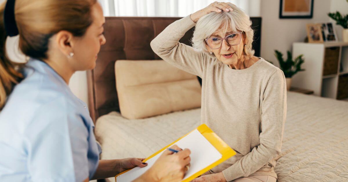 An elderly woman sits on a bed touching her head, speaking to a healthcare worker who is taking notes on a clipboard at St. Mark Village senior living community.