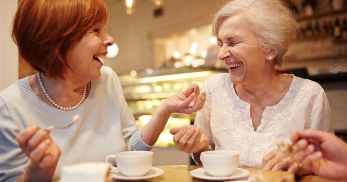 Two elderly women sit at a table in a cafe at St. Mark Village, smiling and laughing together over coffee, enjoying the warmth of senior living.