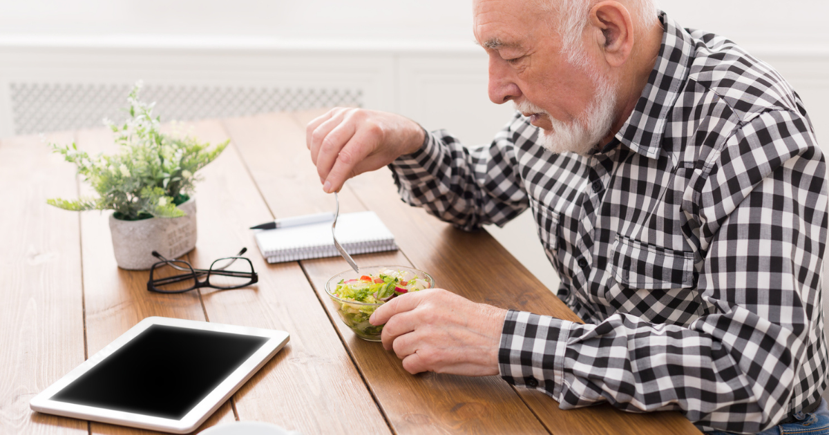 An elderly man in a plaid shirt enjoys salad at a wooden table with a tablet, eyeglasses, and a notebook nearby, capturing the relaxed lifestyle of St. Mark Village senior living.