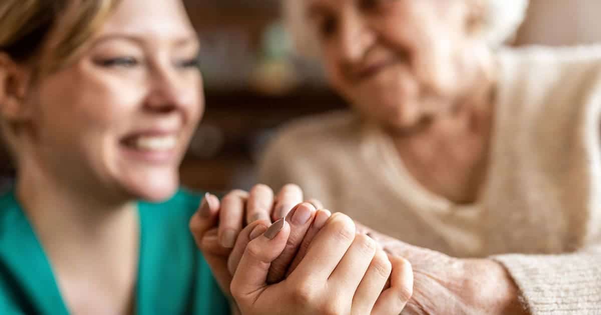 A younger woman smiles while gently holding hands with an older woman, who appears to be offering comfort or support in a warm senior living setting at St. Mark Village.
