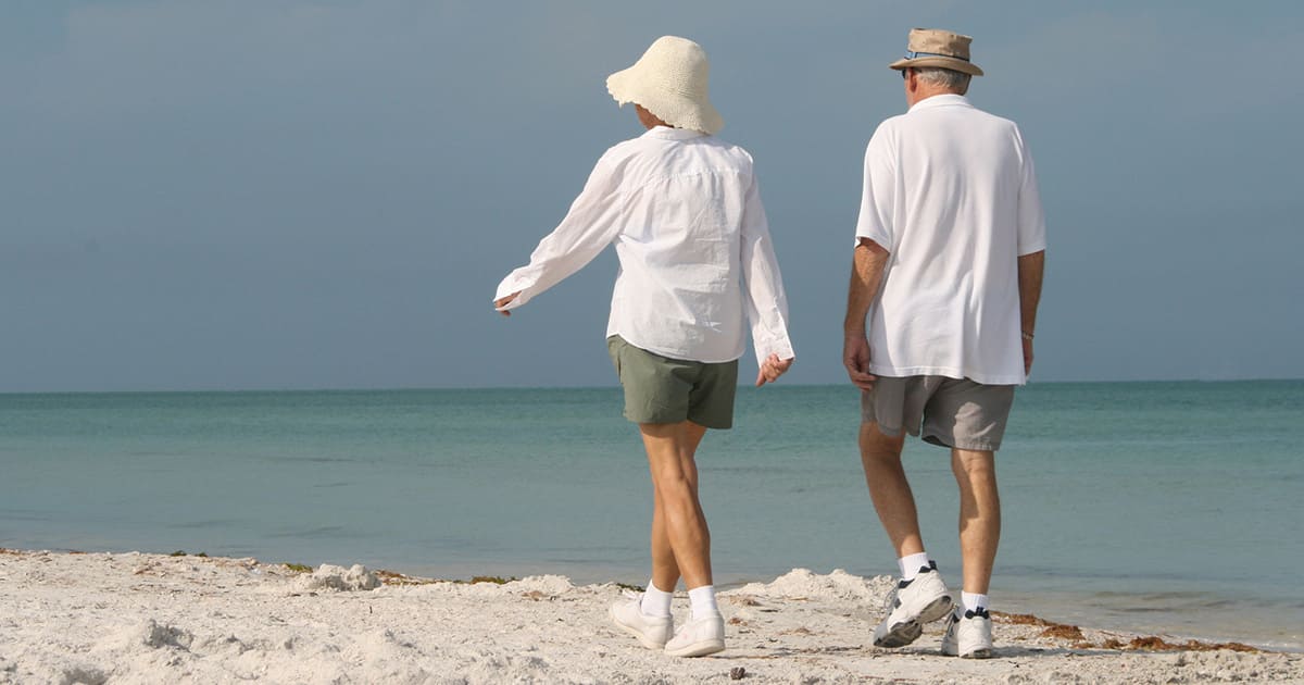 Two older adults enjoy a stroll along a sandy beach near the ocean, both wearing hats and casual summer clothing under a clear sky—embracing the spirit of senior living inspired by St. Mark Village.