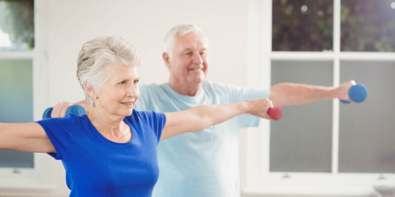 Two older adults stand side by side, lifting small dumbbells with outstretched arms in a bright room at St. Mark Village, enjoying a fitness exercise as part of their vibrant senior living community.