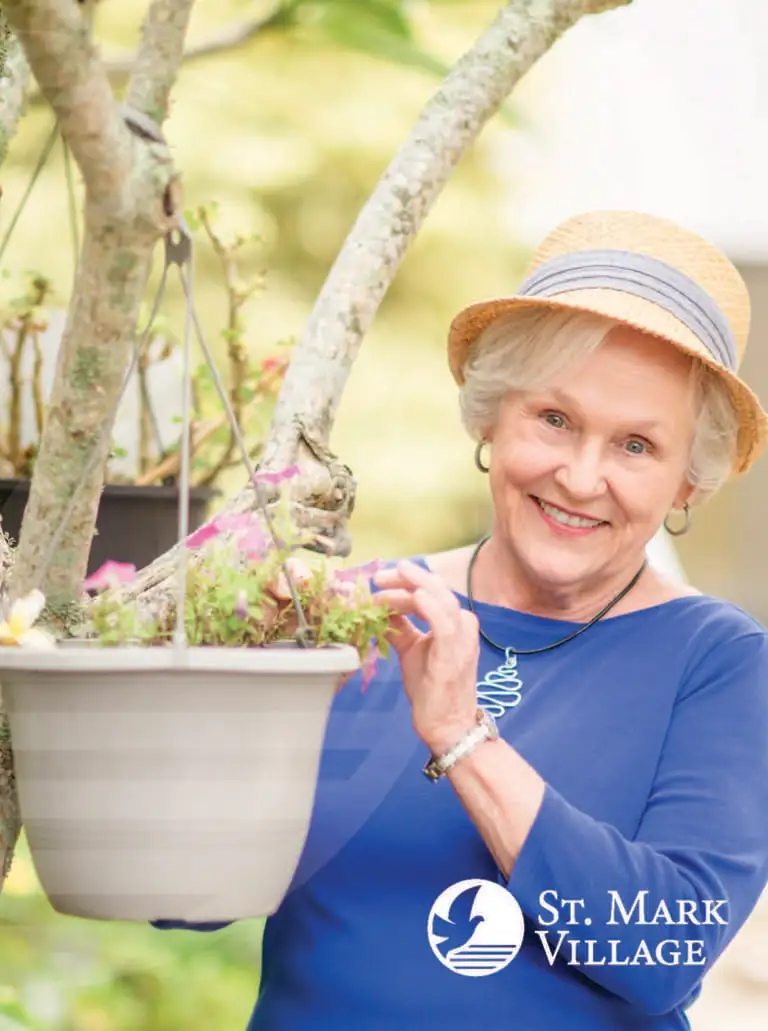 An older woman in a straw hat and blue shirt stands outdoors, smiling as she tends to a hanging potted plant—capturing the joy of senior living at St. Mark Village. The St. Mark Village logo is visible in the corner.