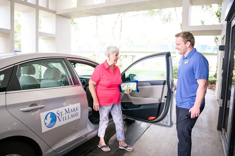 An older woman exits a St. Mark Village senior living car while a staff member in a blue shirt assists her outside a building. An older woman exits a St. Mark Village senior living car while a staff member in a blue shirt assists her outside a building.
