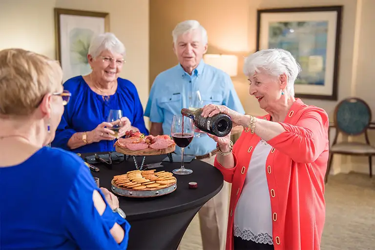 Four older adults stand around a table with snacks at St. Mark Village, as one woman pours wine into a glass, enjoying the vibrant senior living community together. Four older adults stand around a table with snacks at St. Mark Village, as one woman pours wine into a glass, enjoying the vibrant senior living community together.